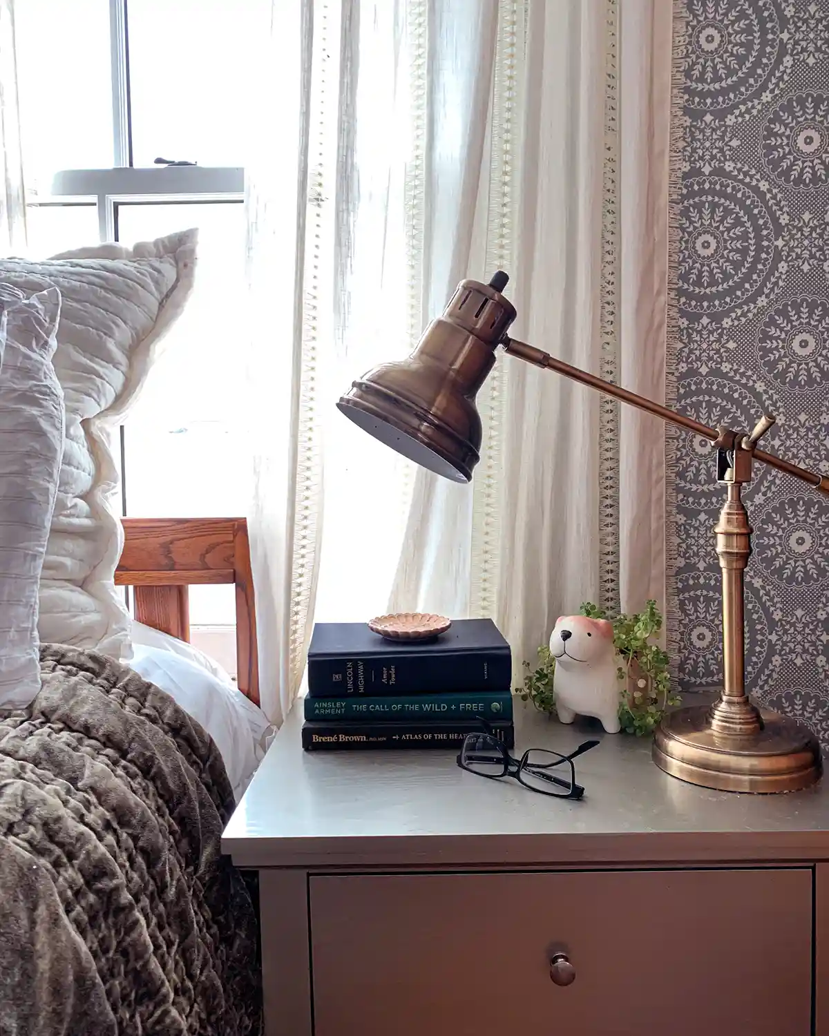 A nightstand sitting beside a bed, clutter free with just a stack of books, ready glasses, lamp, and a plant sitting on top of it